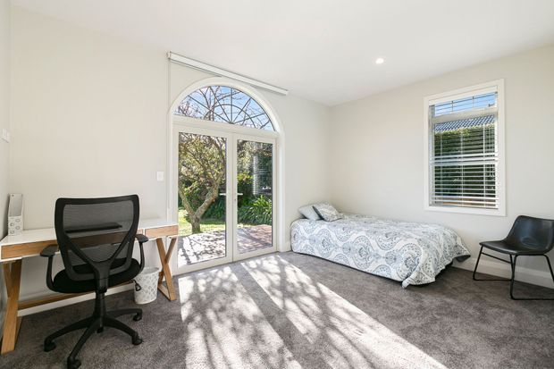 Bright, modest bedroom or home office with a single bed, desk and ergonomic office chair. The room has pale walls, grey carpet, white window and door frames, a glazed arched French door opening to a garden, white plantation-style blinds, and abundant natural light.
