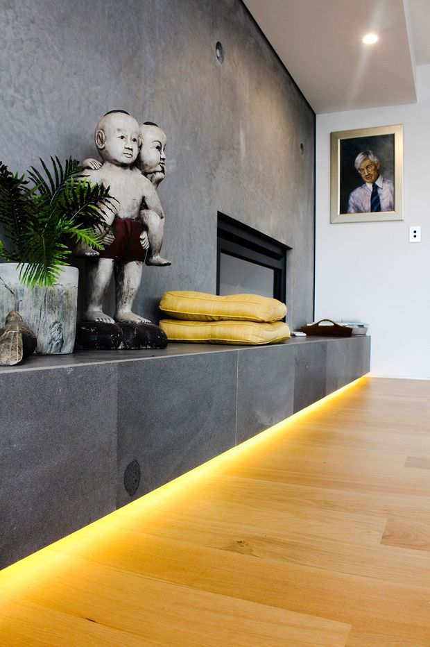 Interior detail of a contemporary living area featuring a dark textured fireplace wall and raised hearth, warm timber flooring with concealed amber underlighting, sculptural figurines, yellow cushions, indoor greenery, and framed artwork.