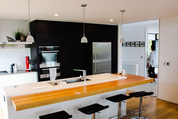 Contemporary open-plan kitchen dominated by a long timber breakfast island with black stools, white cabinetry and benchtops, and a dramatic dark wall of integrated appliances and refrigerator. Pendant lights hang above the island, while a hallway and adjacent living area are visible beyond the kitchen.