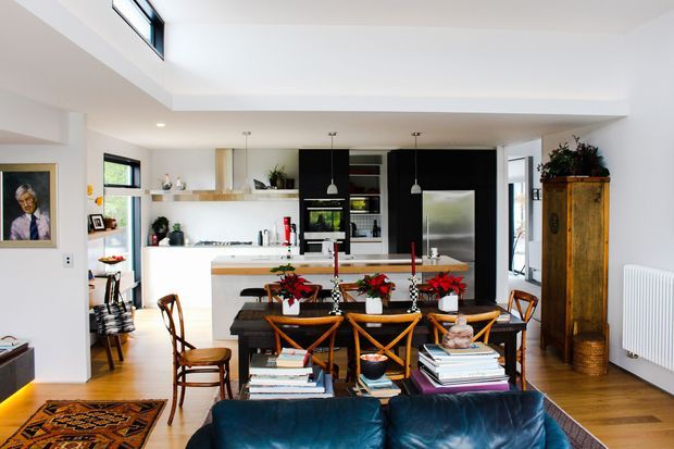 Open-plan interior combining a dining area, kitchen, and living area. The bright space has white walls and ceiling, warm timber flooring, a contemporary black-and-white kitchen with an island, pendant lights, and integrated appliances. A dark wood dining table with cross-back timber chairs sits centrally, while a deep blue sofa occupies the foreground. High-level windows and a raised ceiling section bring in abundant natural light, with a tall traditional timber cabinet adding contrast.