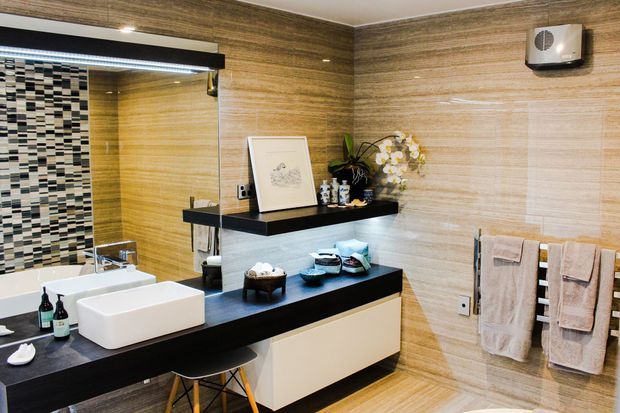 A contemporary bathroom with a wall-mounted floating vanity, white vessel basin, black benchtop and shelving, and warm wood-look tiled walls and floor. A large mirror spans the vanity, while a mosaic-tiled bath or shower area is visible at left. Neutral towels, orchids, and restrained accessories add a refined finish.