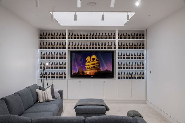 A bright, modern media room with a large grey sectional sofa and ottoman facing a wall-mounted television. The television is integrated into extensive white built-in shelving and cabinetry displaying rows of bottles. A large rectangular ceiling skylight or recessed light well, track lighting, and pale flooring create an airy, clean atmosphere.