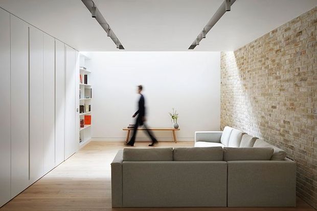Bright, minimalist living room with a large light-grey sectional sofa, pale timber flooring, full-height white built-in cabinetry, open shelving, a simple console table, and an exposed warm brick feature wall. Linear track lighting runs across the white ceiling, creating a clean contemporary interior. A blurred person is walking in the background.