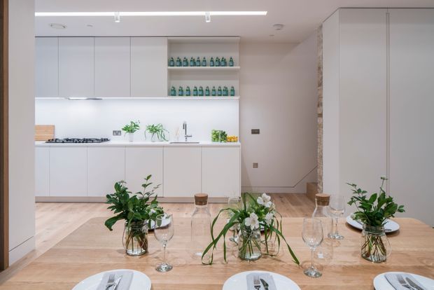 Bright, minimalist open-plan kitchen and dining area with white handleless cabinetry, pale timber flooring and dining table, integrated cooktop and sink, open shelving displaying bottles, and fresh greenery arranged across the dining table. A tall concealed storage wall and recessed stair or passage area are visible at right.