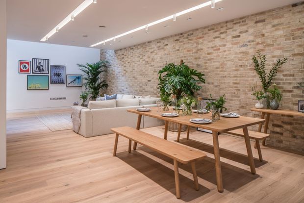 Open-plan interior combining a dining area and living room, with a long timber dining table and benches in the foreground, a pale sectional sofa behind, light timber flooring, exposed warm brick walls, abundant indoor plants, framed artwork, and linear track lighting. The space has a relaxed contemporary-industrial character.