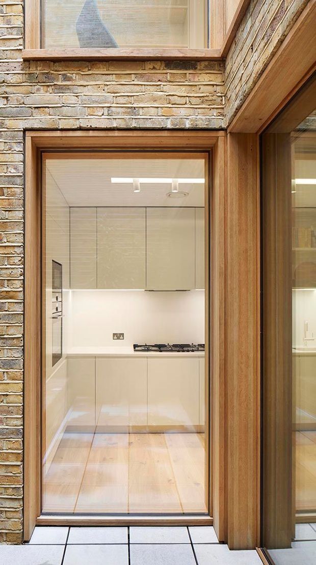 A narrow view into a compact, modern kitchen framed by warm timber glazing and door surrounds. The kitchen features glossy light-coloured handleless cabinetry, integrated under-cabinet lighting, a pale benchtop, gas cooktop, and light timber flooring. The surrounding structure combines exposed light brickwork with timber-framed glazing, creating a strong contrast between the industrial masonry shell and refined contemporary fit-out.