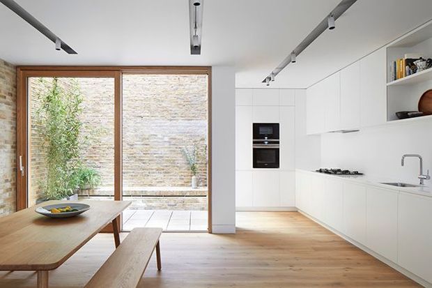 Bright, minimalist open-plan kitchen and dining area with white handleless cabinetry, integrated appliances, a pale stone benchtop, timber flooring, and a timber dining table with bench seating. Full-height timber-framed glazing opens toward a small enclosed courtyard with textured masonry walls. Linear track lighting and exposed brickwork add subtle character.