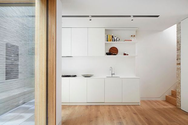 Bright minimalist kitchen and living area viewed through a timber-framed glazed opening. The room features streamlined white handleless cabinetry, open shelving, a slim integrated cooktop and sink, pale timber flooring, recessed linear lighting, and a compact timber stair beside a textured masonry wall. A tiled or stone-lined adjacent area is visible through the glazing on the left.