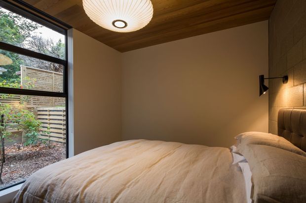 Compact bedroom with a low-profile bed, cream upholstered headboard, and white textured bedding. The room features a warm timber-lined ceiling, soft off-white walls, a large black-framed window overlooking a planted outdoor area, and discreet wall-mounted lighting. The overall design is warm, calm, and contemporary with natural materials.