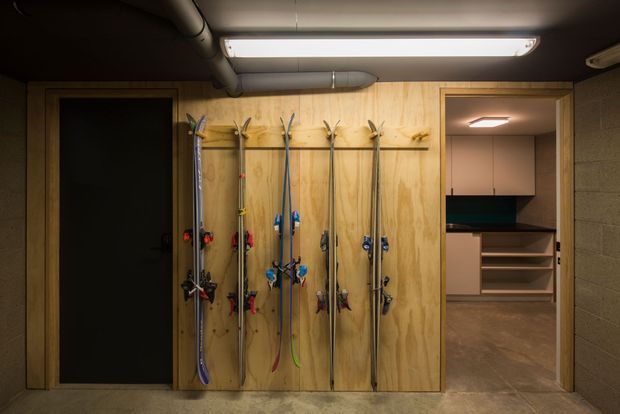 A compact ski storage and equipment area with several pairs of skis mounted vertically on a plywood wall panel. The space has exposed ceiling services, an elongated fluorescent light, dark concrete-block walls, and a doorway leading to built-in shelving and cabinetry.