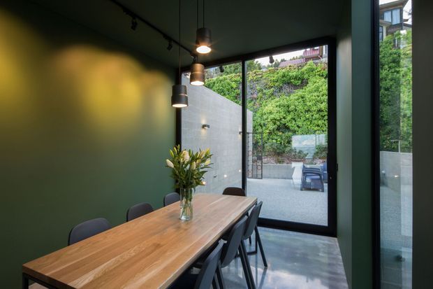 A moody, contemporary dining room with a long solid timber dining table, dark upholstered chairs, deep green walls and ceiling, black pendant lights, and polished concrete flooring. Full-height glazed doors open onto a small paved courtyard with greenery and an outdoor seating area.
