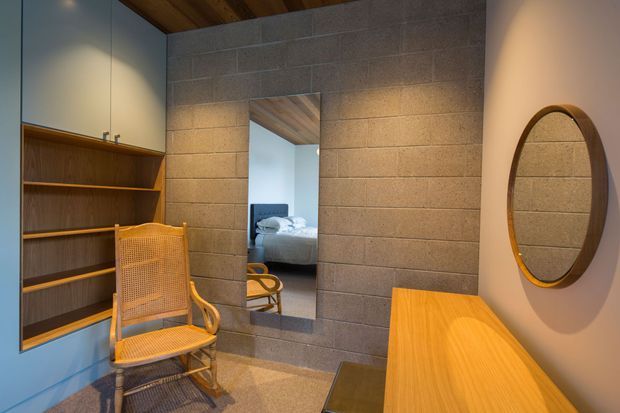 A warm, compact bedroom dressing area with a built-in timber shelving unit and pale cabinetry, a woven cane lounge chair, a timber console, and a round timber-framed wall mirror. Grey masonry block walls and a timber-lined ceiling frame a full-height opening that reveals a bed in the adjoining bedroom.