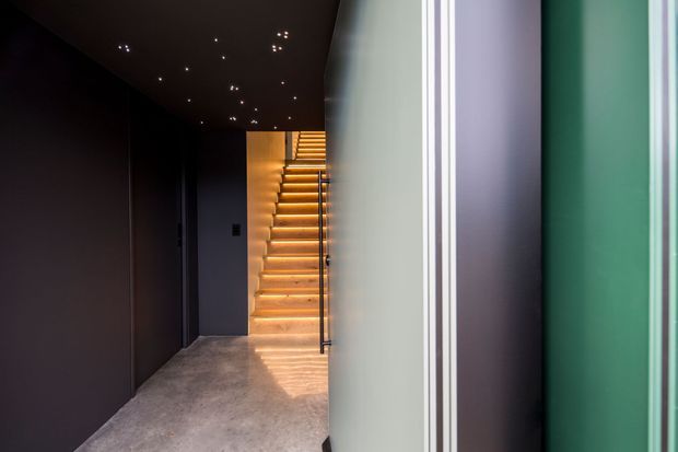 A moody contemporary hallway or entry passage with dark painted walls and ceiling, a polished concrete floor, and a warmly illuminated staircase at the rear. Slim recessed ceiling spotlights and a dark metal stair handrail create a dramatic, minimalist atmosphere. A partially visible green-toned doorway or wall frames the right side.
