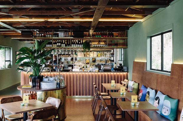 A compact, warmly lit bar and café interior with a long ribbed timber-toned bar front, extensive open shelving of bottles and glassware, and a dark exposed timber ceiling with visible structural framing. Green-painted walls, large black-framed windows, polished flooring, indoor tropical plants, and small timber tables with mixed seating create a lively hospitality setting. A built-in upholstered bench with colourful illustrated cushions runs along one wall.