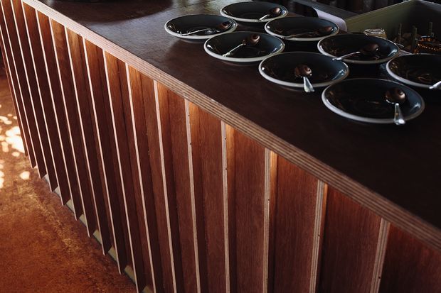 Close-up view of a dark timber bar or counter with vertically slatted wood panelling and a layered timber countertop edge. Several black bowls and serving dishes sit on the surface. Warm, directional lighting highlights the rich wood grain and textured carpet below.