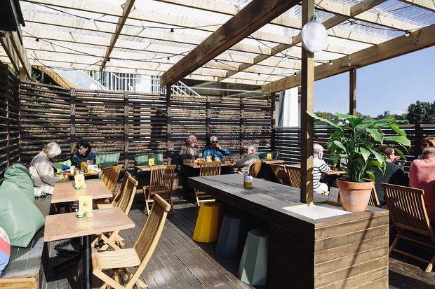 An open-air restaurant or café terrace with timber decking, pergola-style overhead structure, horizontal timber privacy screens, communal and individual dining tables, and built-in bench seating. Large potted tropical plants and simple pendant lights add warmth to the sunny outdoor setting.
