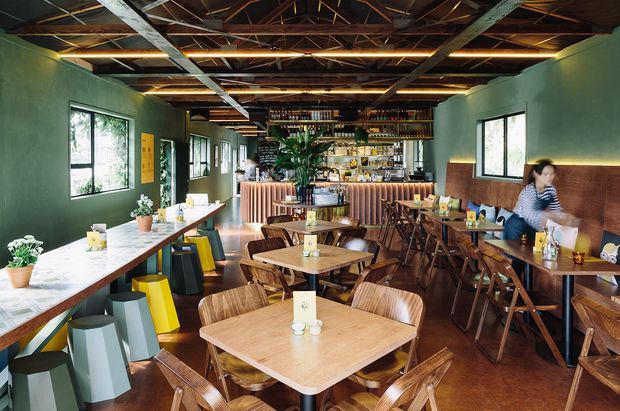 A warmly lit, industrial-style restaurant interior with rows of timber dining tables and chairs, colourful geometric stools, dark green walls, exposed timber roof trusses and services, and a bar at the rear with ribbed timber cladding and open shelving. Large windows bring in natural light, while plants and table settings add a welcoming atmosphere.