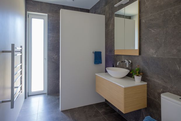 Contemporary bathroom with dark stone-look tiled walls and floor, a floating light-wood vanity with a white vessel basin, wall mirror, white toilet, heated towel rail, and a narrow frosted-glass window providing natural light.