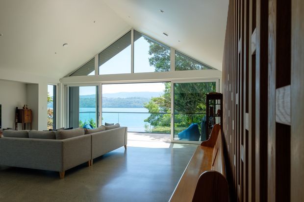 A bright contemporary living room opening onto a waterfront view. The room features a large grey sectional sofa, polished concrete flooring, extensive floor-to-ceiling glazing with a dramatic triangular clerestory, and a timber slatted feature wall with integrated shelving and bench seating. Trees, blue water, and distant hills are visible beyond the glazing.