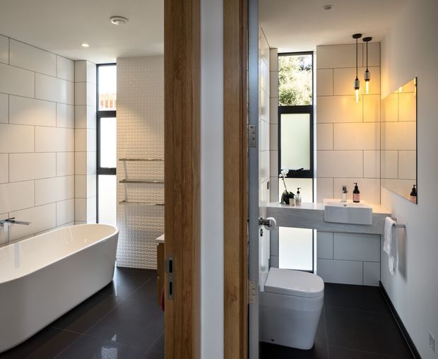 A contemporary bathroom suite viewed through an open timber-framed doorway, showing a freestanding white bathtub in the left area and a separate vanity and toilet area on the right. The room has large white rectangular wall tiles, dark tiled flooring, frameless or dark-framed windows, a floating grey vanity with integrated storage, a white vessel basin, wall-mounted taps, and pendant lighting. The image presents two connected bathroom zones, with no bedroom visible.