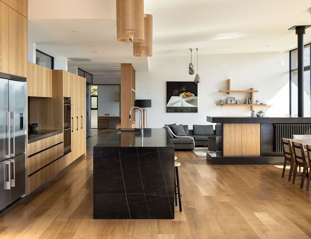 A bright, open-plan contemporary kitchen and living area with extensive light timber cabinetry, a large dark stone island, timber flooring, and black-framed glazing. The kitchen includes integrated appliances and island seating, while the adjoining living zone has a sofa, wall-mounted shelving, and a dark kitchen or bar counter separating it from the dining area.