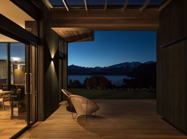 A dramatic covered outdoor terrace or veranda opening toward a lake and mountain landscape at dusk. The space features warm timber decking, dark vertical timber wall cladding, a timber-lined ceiling with exposed beams and integrated lighting, extensive glass sliding doors, and a woven lounge chair. A warmly lit interior living area is visible through the glazing on the left.