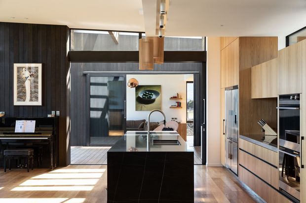 A bright, contemporary open-plan kitchen viewed straight on, with a large dark stone island and integrated sink in the foreground. Full-height timber cabinetry with built-in refrigerator and ovens lines the right wall, while dark vertical timber wall panelling and a piano occupy the left side. Glazed doors and windows open toward an outdoor courtyard, and a sculptural timber pendant hangs above the island. The room combines dark feature finishes with warm timber and abundant natural light.