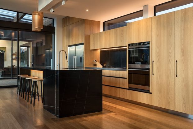 Contemporary open-plan kitchen with a large black stone island and integrated breakfast bar, light timber cabinetry, black tiled splashback, built-in stainless-steel appliances, and warm timber flooring. Dark-framed glazing and a connected dining or living area are visible beyond the kitchen. The space has clean linear detailing, warm lighting, and a refined, high-contrast material palette.