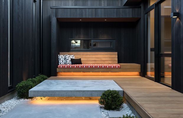 A compact contemporary courtyard or outdoor lounge area with a built-in timber bench beneath a dark vertical-panelled facade. The bench has patterned cushions and warm integrated under-seat lighting, with a large pale concrete platform in front, timber decking to the right, black-framed glazing, minimalist wall lighting, white gravel, and clipped shrubs.