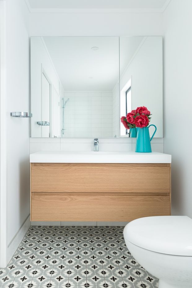 Bright, compact contemporary bathroom viewed straight on. A wall-mounted light timber vanity with two wide drawers sits beneath a large mirrored cabinet, paired with a white integrated basin and chrome tapware. White tiled shower walls are reflected in the mirror, while patterned geometric floor tiles add visual interest. A white toilet occupies the lower right, and a turquoise vase with vivid red flowers provides a small colour accent.