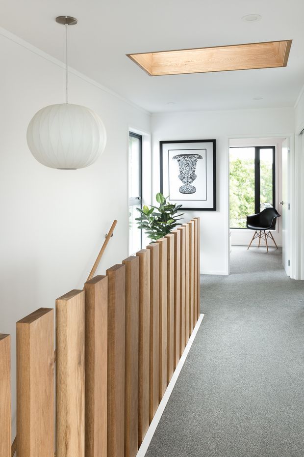 Bright contemporary upper-level hallway and stair landing with white walls, grey carpet, a sculptural vertical timber stair balustrade, and a large recessed timber-lined ceiling lightwell. A spherical pendant light, framed artwork, indoor plant, and glimpses of an adjacent room with a black chair are visible.