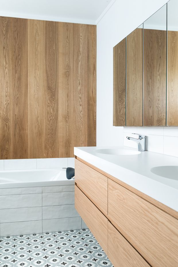 Bright contemporary bathroom featuring a long white double vanity with light timber-grain cabinetry, integrated basins, chrome tapware, and mirrored wall cabinets. The room includes a bathtub with pale grey wall tiling, a vertical timber-look feature wall, and patterned encaustic-style floor tiles in white, grey, and black.