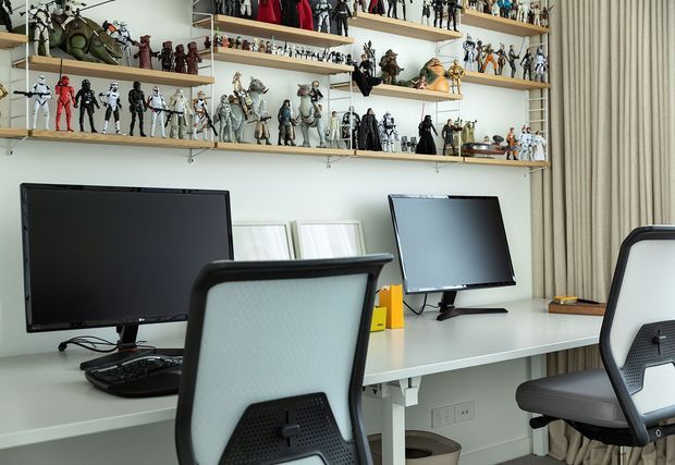 A bright, contemporary home office or studio workspace with a long shared white desk, multiple large computer monitors, ergonomic office chairs, and extensive wall-mounted timber shelving. The shelves are densely arranged with collectible figurines and models, creating a highly personalised display. Neutral walls, light furnishings, and full-height beige curtains give the room a clean, organised appearance.