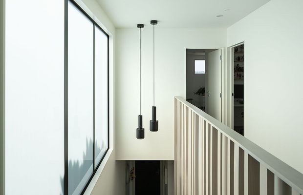 A bright minimalist stair landing with a white-painted balustrade, large black-framed glazing, and two simple black pendant lights. The landing overlooks a lower level and connects to a hallway, with a built-in storage or wardrobe area visible through an open door.