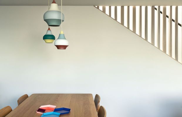 Minimal dining area with a large warm timber dining table and wooden chairs beneath three sculptural pendant lights in muted teal, cream, yellow, and red. A white stairwell wall and open vertical timber balustrade create a strong diagonal architectural feature in the background. The space is bright and uncluttered, with a contemporary, playful character.