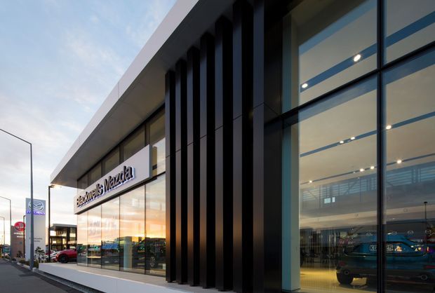 Exterior photograph of a contemporary commercial building, likely a retail or automotive showroom. The facade combines extensive floor-to-ceiling glazing with dark vertical fins, a projecting light-coloured roof edge, and illuminated interior display areas visible through the windows. Street signage, parked vehicles, and neighbouring commercial elements appear in the background.