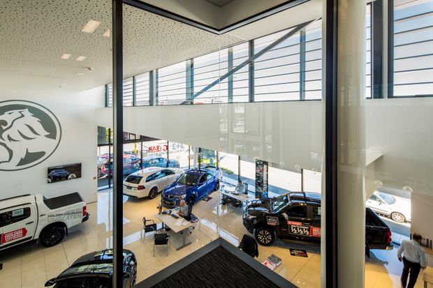 A bright, double-height automotive showroom viewed from an upper-level glazed interior walkway or office. Floor-to-ceiling glass partitions overlook polished vehicle display areas with cars, customer seating, and desks. Large horizontal clerestory-style windows, black metal framing, a suspended acoustic tile ceiling, and a prominent circular automotive logo define the commercial space.