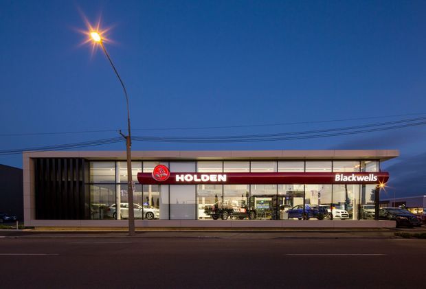 A modern single-storey Holden car dealership photographed at dusk from across the street. The broad glazed showroom frontage is framed by dark vertical fins and a simple low horizontal roofline, with illuminated Holden and Blackwells signage, vehicles visible inside, street lighting, and overhead utility wires.