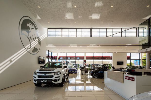 Bright, spacious automotive dealership showroom and reception area with glossy tiled flooring, high ceilings, extensive glazing, vehicle displays, and a white reception desk. A large circular wall graphic and elevated mezzanine-like upper level are visible, with strong daylight entering through clerestory windows.