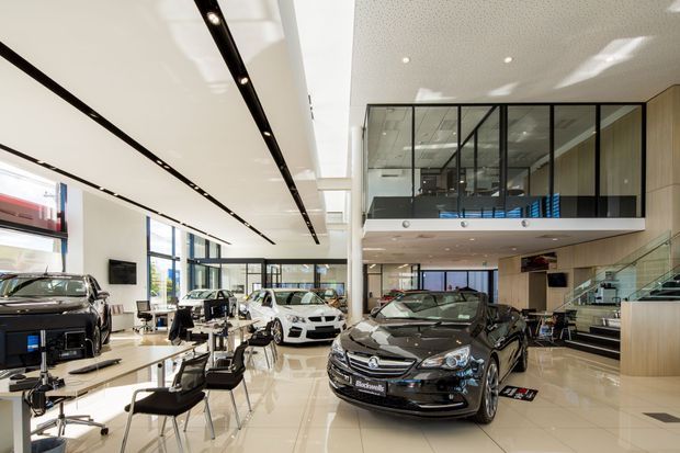 Bright, double-height automotive showroom with several display vehicles arranged on a glossy light-coloured floor. The space features extensive floor-to-ceiling glazing, a dramatic linear ceiling lighting system, a glass-fronted mezzanine or upper-level office, a reception or consultation area, and a staircase with glass balustrading. The architecture is sleek, commercial, and highly polished.
