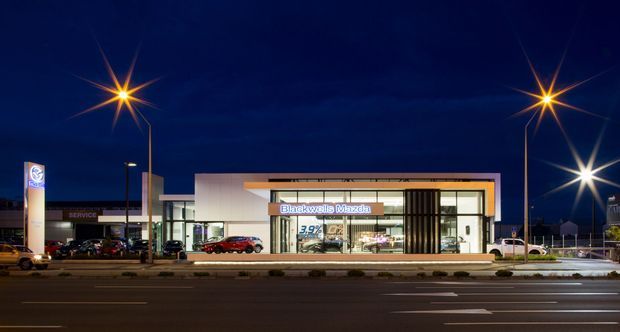Night-time exterior photograph of a modern Mazda automotive dealership viewed across a multi-lane road. The low-rise commercial building has a broad glazed showroom frontage, pale rendered walls, a projecting horizontal canopy with warm timber-toned trim, and prominent Mazda and dealership signage. Exterior streetlights and illuminated showroom interiors are visible.