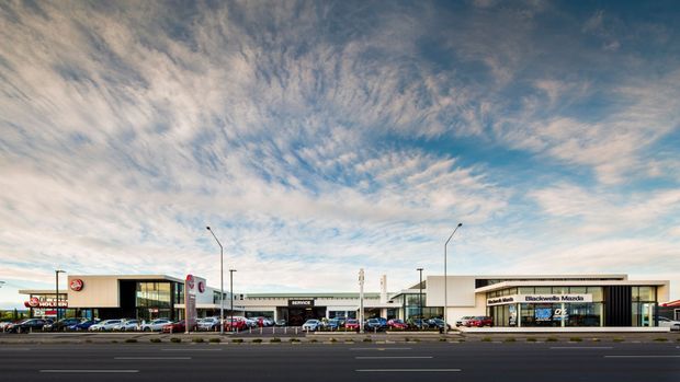 Wide exterior photograph of a contemporary single-storey commercial retail and automotive complex viewed across a multi-lane road. The development has a long, low horizontal form with flat roofs, pale rendered or metal-clad facades, extensive storefront glazing, prominent business signage, parking, and street lighting. A dramatic cloud-filled sky occupies much of the upper frame.