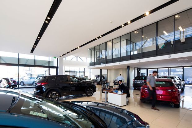 A bright, spacious multi-level automotive showroom with vehicles displayed across a polished tiled floor. Full-height glazing creates an airy frontage, while a black-framed glass mezzanine and linear recessed track lighting define the contemporary interior. A compact reception or sales desk sits among the cars.
