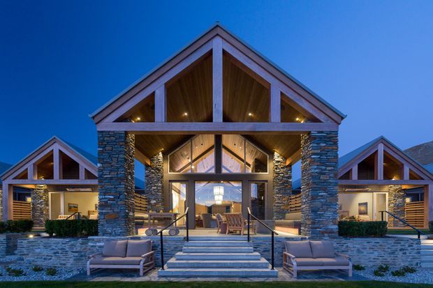 Symmetrical lodge-style building exterior photographed at dusk, featuring prominent front-facing gabled roofs, stacked-stone columns and walls, warm timber soffits and exposed structural framing, expansive glazed entry doors, broad stone steps, and outdoor lounge seating. Illuminated interiors and architectural lighting create a welcoming alpine or resort-like appearance.