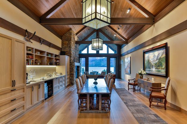 A warm, expansive dining room with a long timber dining table and upholstered chairs, extensive built-in timber cabinetry and display shelving, a stone-clad fireplace or structural column, timber floors, and dramatic exposed timber roof beams. Large glazed doors and clerestory windows open the room toward a scenic landscape. Two substantial geometric pendant lights hang from the vaulted ceiling, while framed artwork and occasional seating furnish the perimeter.