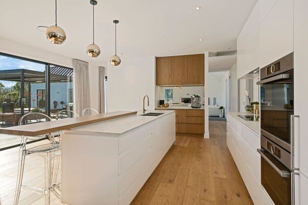 Bright contemporary open-plan kitchen with a large white island, integrated cabinetry, timber flooring, timber overhead joinery, built-in stainless-steel appliances, and a dining area beside expansive glazing. Three spherical pendant lights hang above the island, while a covered outdoor area is visible beyond the glazing.