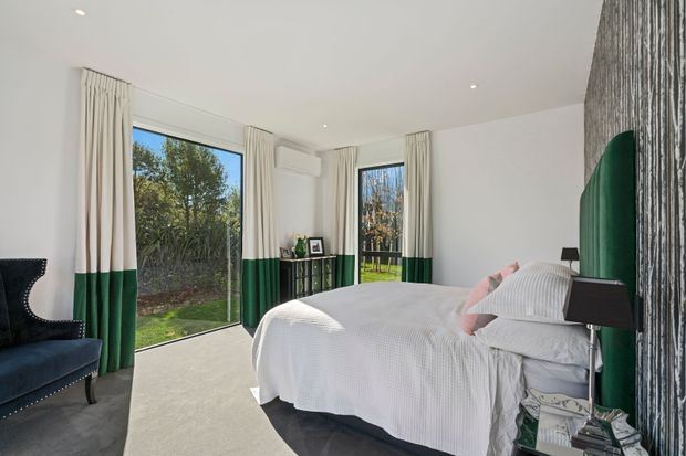 Bright contemporary bedroom with a large bed dressed in white bedding, green upholstered headboard, dark bedside tables, and floor-to-ceiling windows with white-and-deep-green curtains. The room has pale carpet, white walls and ceiling, recessed lighting, and a textured dark feature wall. Garden greenery is visible outside the glazing.
