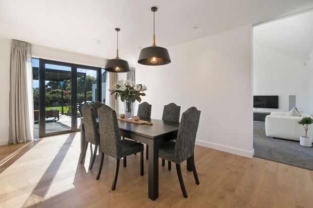 Bright, contemporary dining area with a dark rectangular dining table and six upholstered chairs, set on light timber flooring. Three black pendant lights hang above the table. Full-height glazed doors open to an outdoor area, while an adjacent living room with a white sofa is visible through a wide opening.