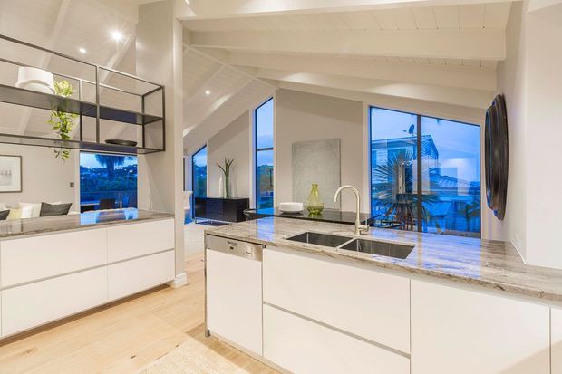 Bright contemporary open-plan kitchen with a large white island, integrated sink and dishwasher, pale stone benchtops, light timber flooring, black-framed open shelving, and extensive glazing opening to an outdoor deck or balcony. The room has a high exposed-beam ceiling and views toward a built-up coastal or suburban setting.