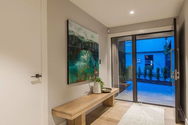 A refined contemporary entryway with a warm timber console bench, abstract artwork, ceramic vase with greenery, and a textured rug. Full-height glazed sliding doors open to a paved outdoor courtyard, while recessed ceiling lighting and neutral walls create a calm, inviting atmosphere.